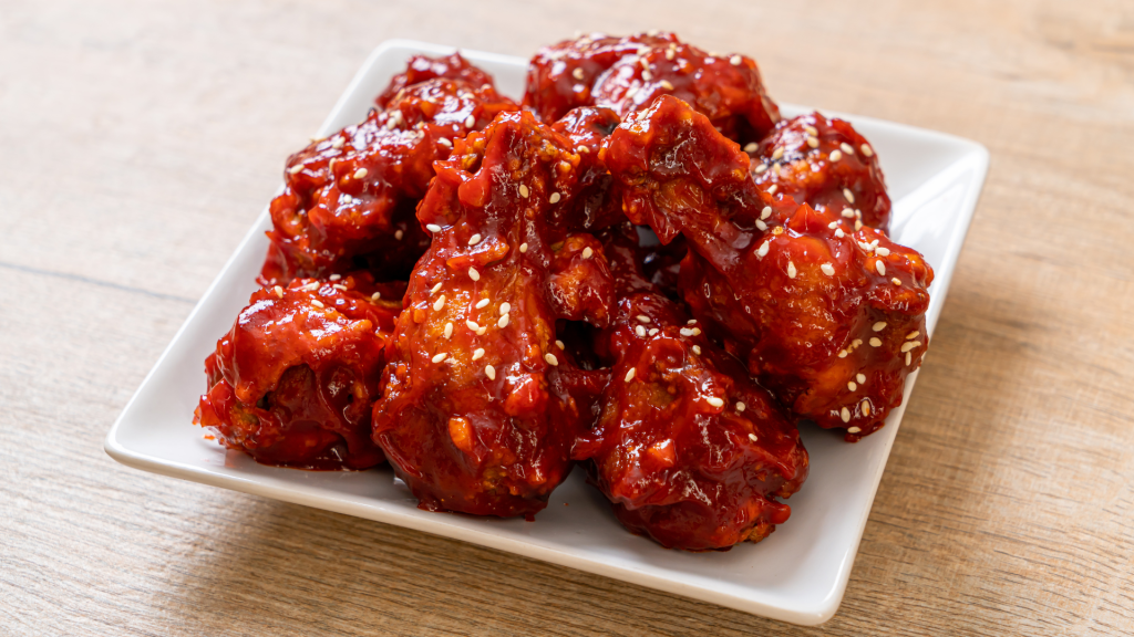 A plate of crispy Korean fried chicken wings, golden-brown and glistening with a thick, sticky gochujang glaze. The sauce has a deep red hue, hinting at its spicy-sweet flavor. Garnished with sesame seeds and finely chopped green onions, the chicken is served on a rustic ceramic plate with a side of pickled radish and a small bowl of extra sauce. The Ultimate Guide to Catering a Party at Home: Essential Tips and 10 Best Ideas for Melbourne Hosts